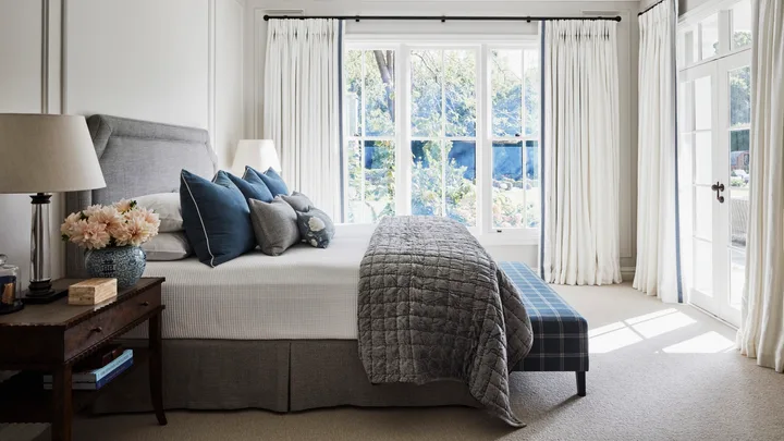A light-filled bedroom with a cream coloured carpet, white walls, white curtains open to reveal a leafy backyard. A grey bed is in the centre of the room, with a grey quilt folded over the end of the white bed sheets and plenty of white and navy pillows on top. A bedside table is beside the bed, with a lamp, blue and white vase with peach toned dahlias inside and a wooden box.