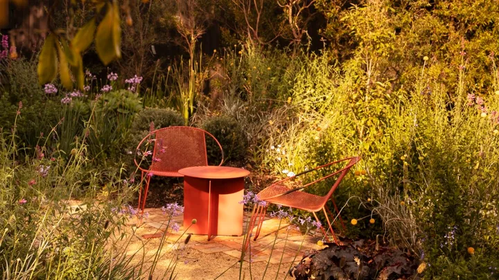 A cottage garden, with two red outdoor chairs, beside a red outdoor side table, positioned on a brick and gravel path, with garden beds on either side. Leaves from a tree are in the foreground.