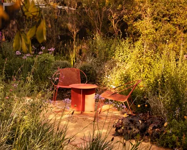 A cottage garden, with two red outdoor chairs, beside a red outdoor side table, positioned on a brick and gravel path, with garden beds on either side. Leaves from a tree are in the foreground.