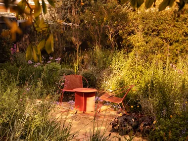 A cottage garden, with two red outdoor chairs, beside a red outdoor side table, positioned on a brick and gravel path, with garden beds on either side. Leaves from a tree are in the foreground.