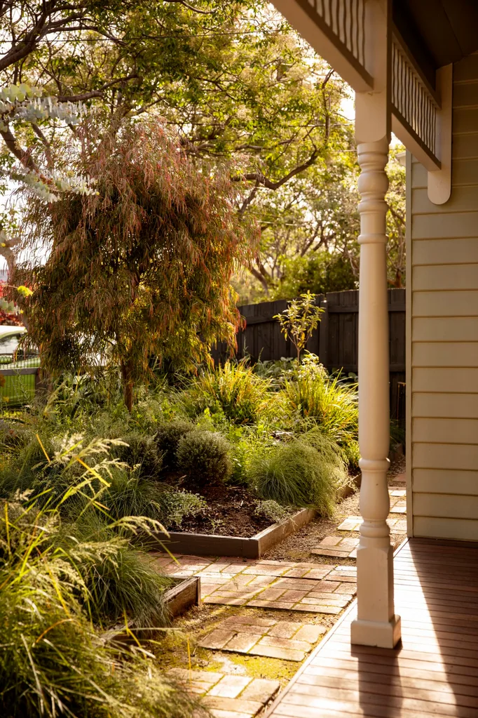 The front garden of a grey coloured cottage. The porch of the cottage is visible to the right, while the rest of the photo focuses on the brick and gravel pathway connecting different garden beds that are filled with foliage.