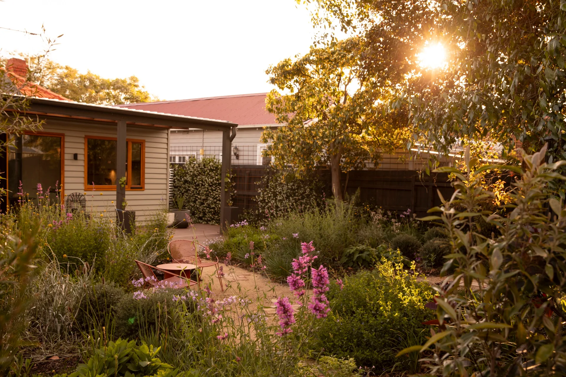 Sunlight softly filteres through a tree, casting the backyard of this cottage in a golden glow. The cottage garden features winding gravel and brick paths, two red outdoor chairs beside wild garden beds overflowing with foliage and colourful flowers.