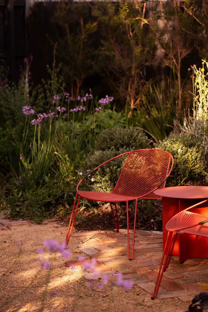 A close up photo of two red outdoor chairs, beside a red outdoor side table. They are positioned on a brick and gravel path, with garden beds on either side.
