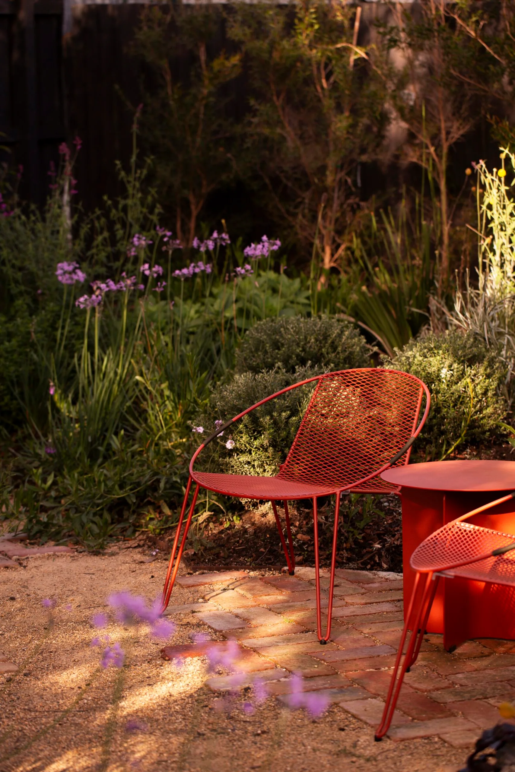 A close up photo of two red outdoor chairs, beside a red outdoor side table. They are positioned on a brick and gravel path, with garden beds on either side.