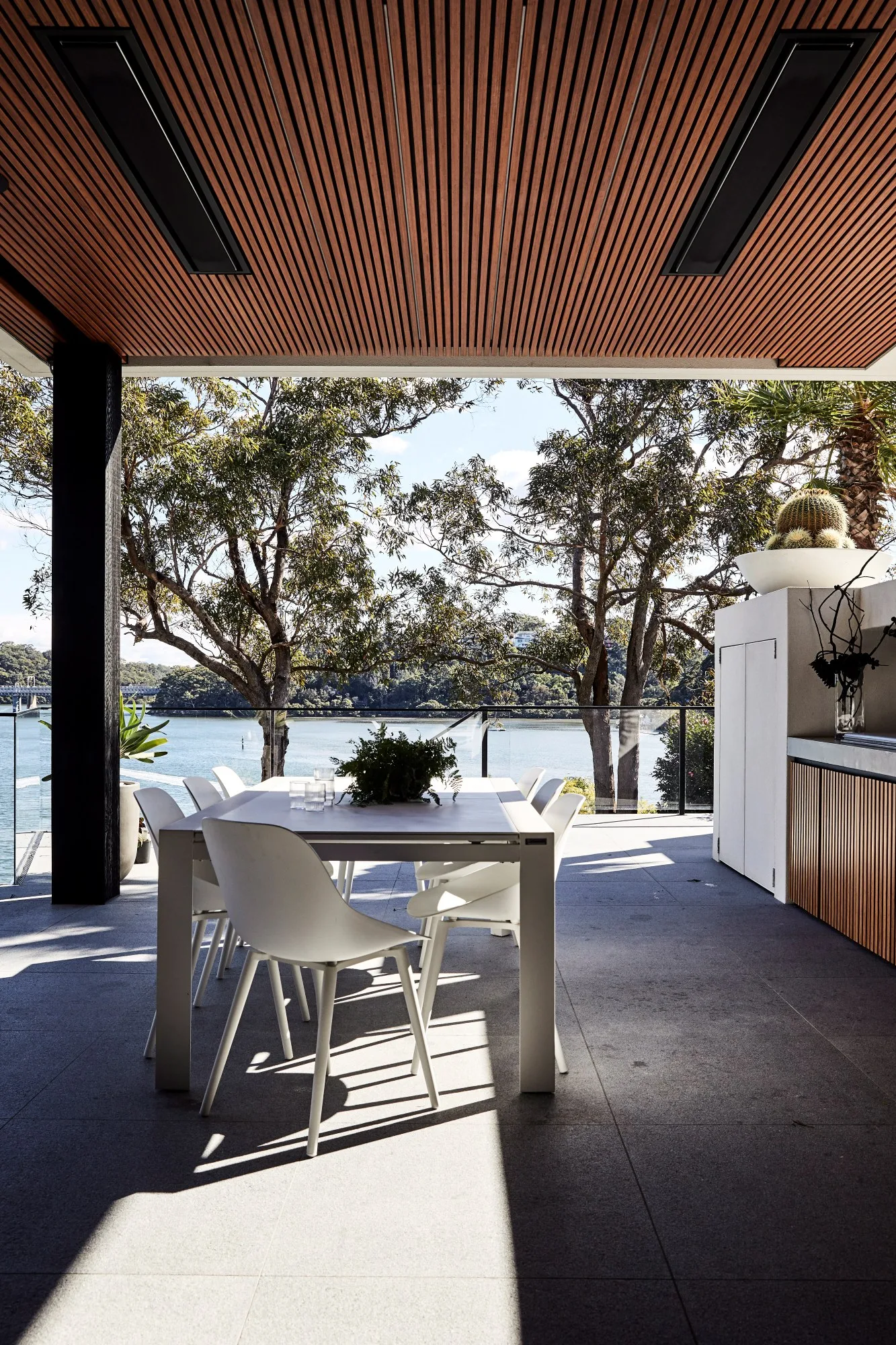Covered outdoor dining area with white chairs, table, and a view of trees and water through large windows.