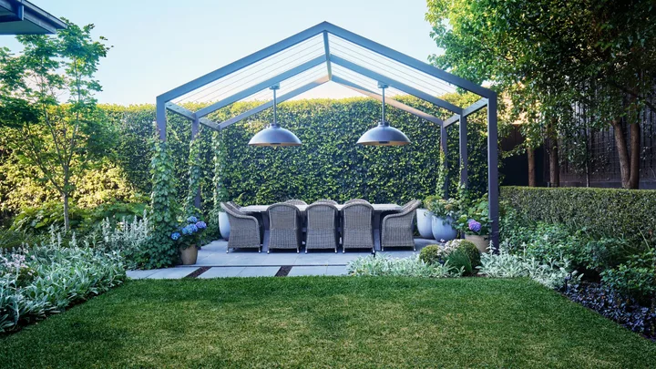 Outdoor dining area beneath a steel pergola surrounded by lush hedges and soft garden planting.