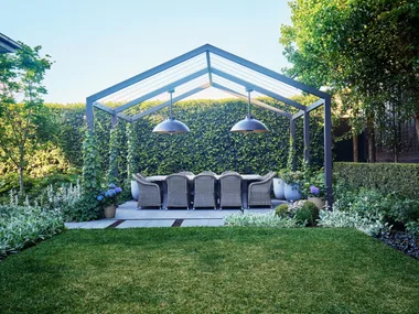 Outdoor dining area beneath a steel pergola surrounded by lush hedges and soft garden planting.