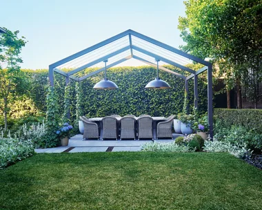 Outdoor dining area beneath a steel pergola surrounded by lush hedges and soft garden planting.