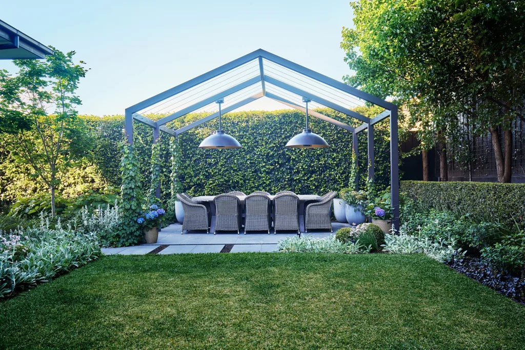 Outdoor dining area beneath a steel pergola surrounded by lush hedges and soft garden planting.