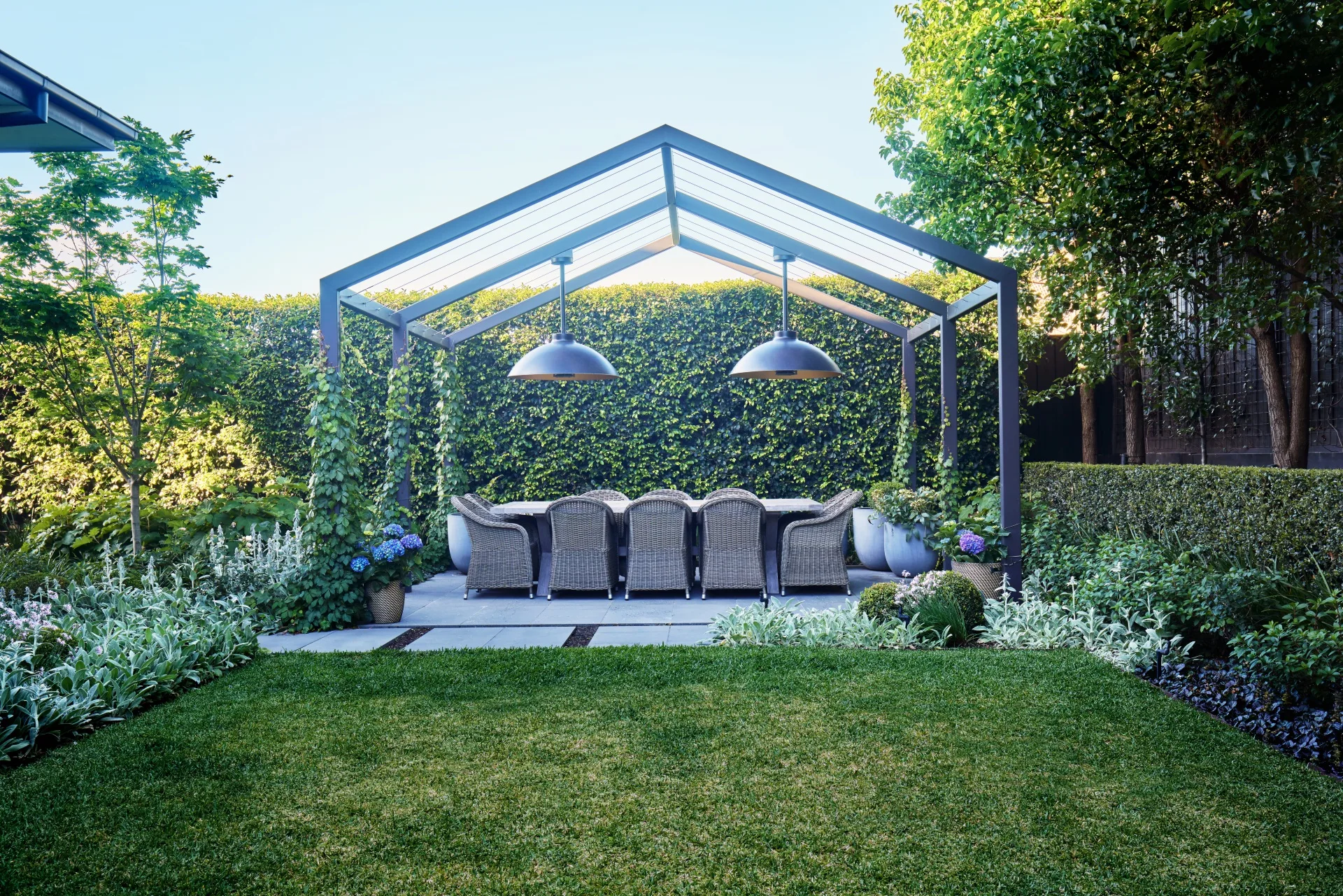 Outdoor dining area beneath a steel pergola surrounded by lush hedges and soft garden planting.