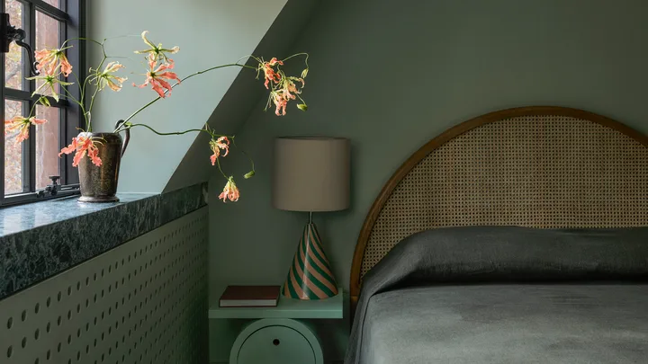A cozy colour-drenched on-trend bedroom in 2026 with green walls, window seat with flowers, lamp on side table, and arched headboard.