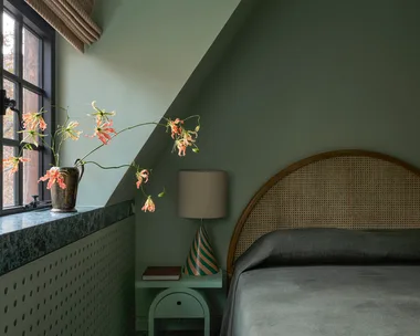 A cozy colour-drenched on-trend bedroom in 2026 with green walls, window seat with flowers, lamp on side table, and arched headboard.