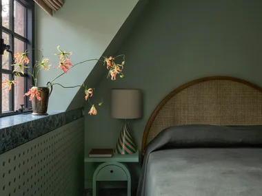 A cozy colour-drenched on-trend bedroom in 2026 with green walls, window seat with flowers, lamp on side table, and arched headboard.