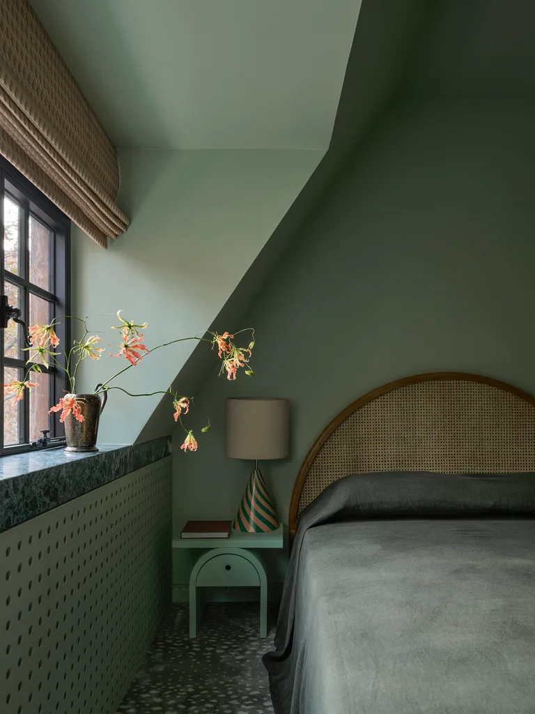A cozy colour-drenched on-trend bedroom in 2026 with green walls, window seat with flowers, lamp on side table, and arched headboard.