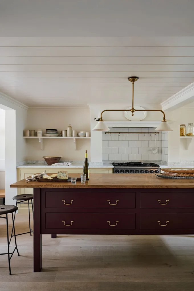 Kitchen interior in 2026 with freestanding dark timber island, wooden countertop, cheese, baguette, wine bottle, and vintage lighting fixture.