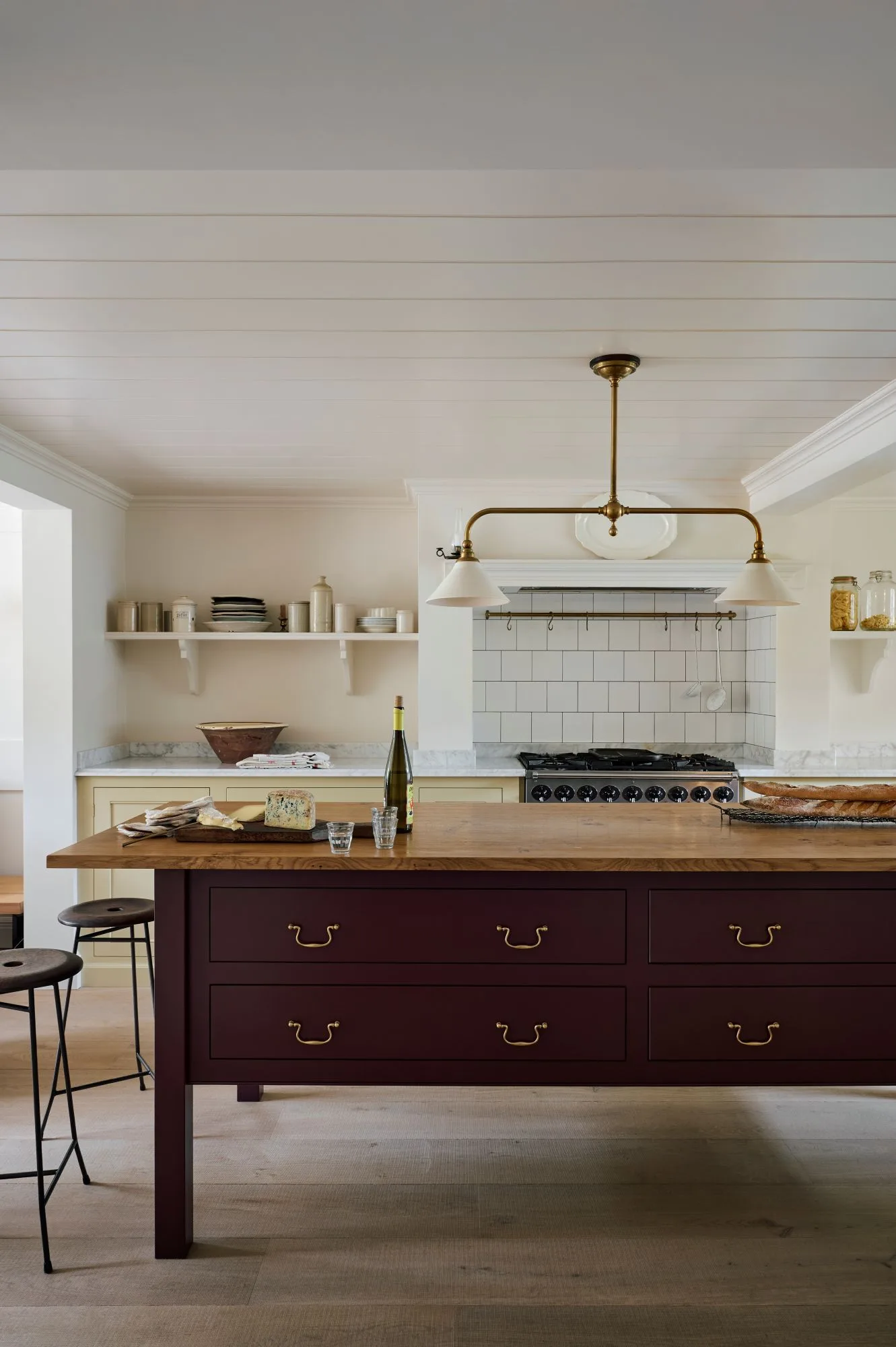 Kitchen interior in 2026 with freestanding dark timber island, wooden countertop, cheese, baguette, wine bottle, and vintage lighting fixture.