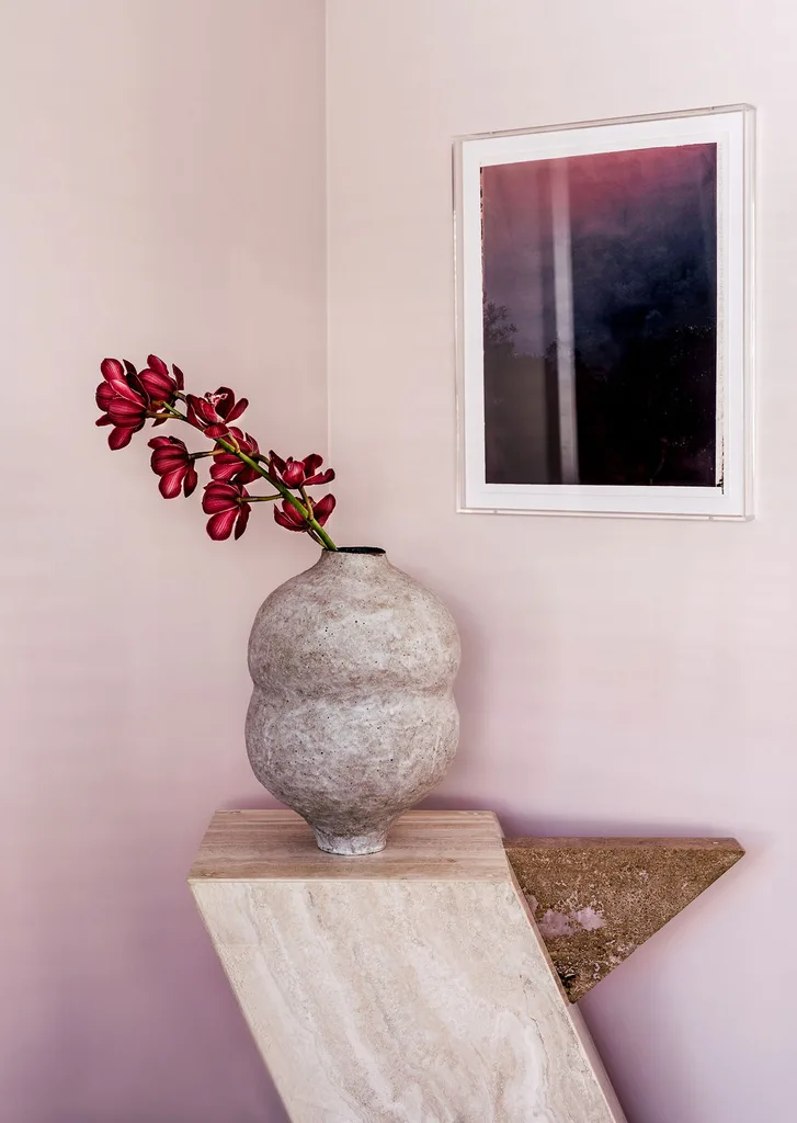 A ceramic vase with a pink floral branch on a marble plinth against a pale pink wall with a deep pink and purple artwork