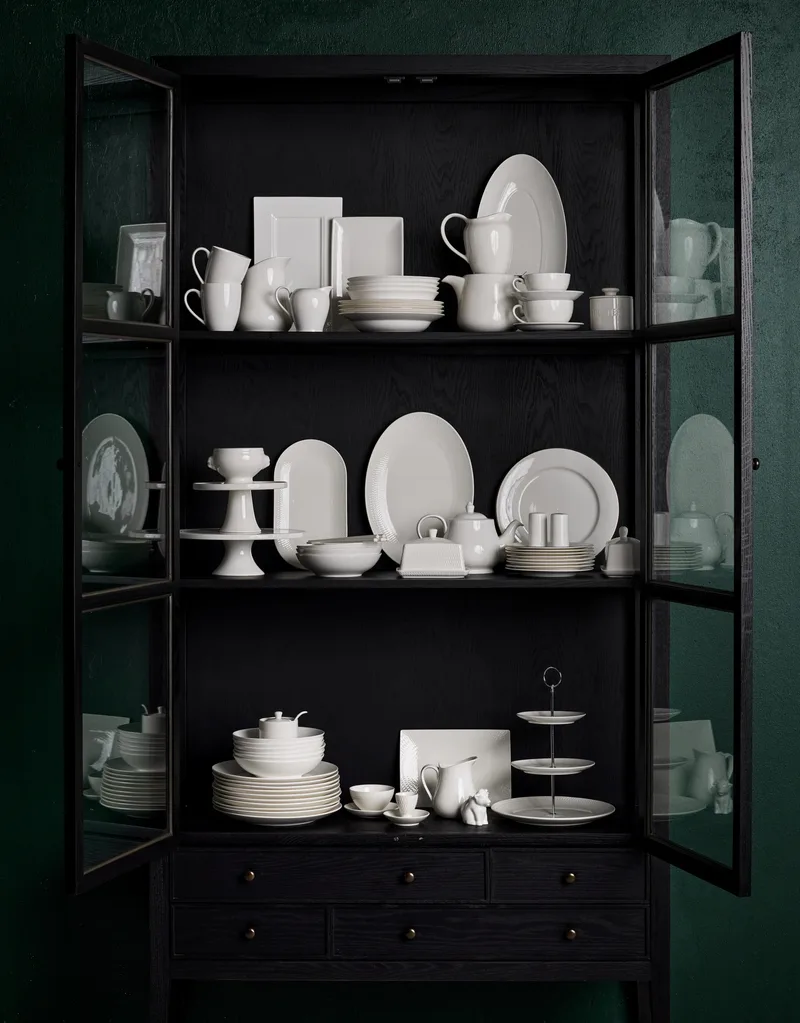 An open black cabinet with shelves of white dinner sets, tea seats and other tableware. All the pieces are white and ceramic.