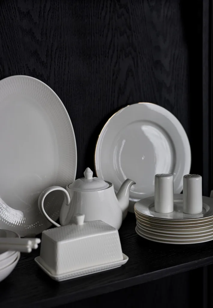A black cabinet shelf with two round white plates, a white teapot, a white butter dish and a white stack of small bread plates are all displayed in the cabinet shelf.