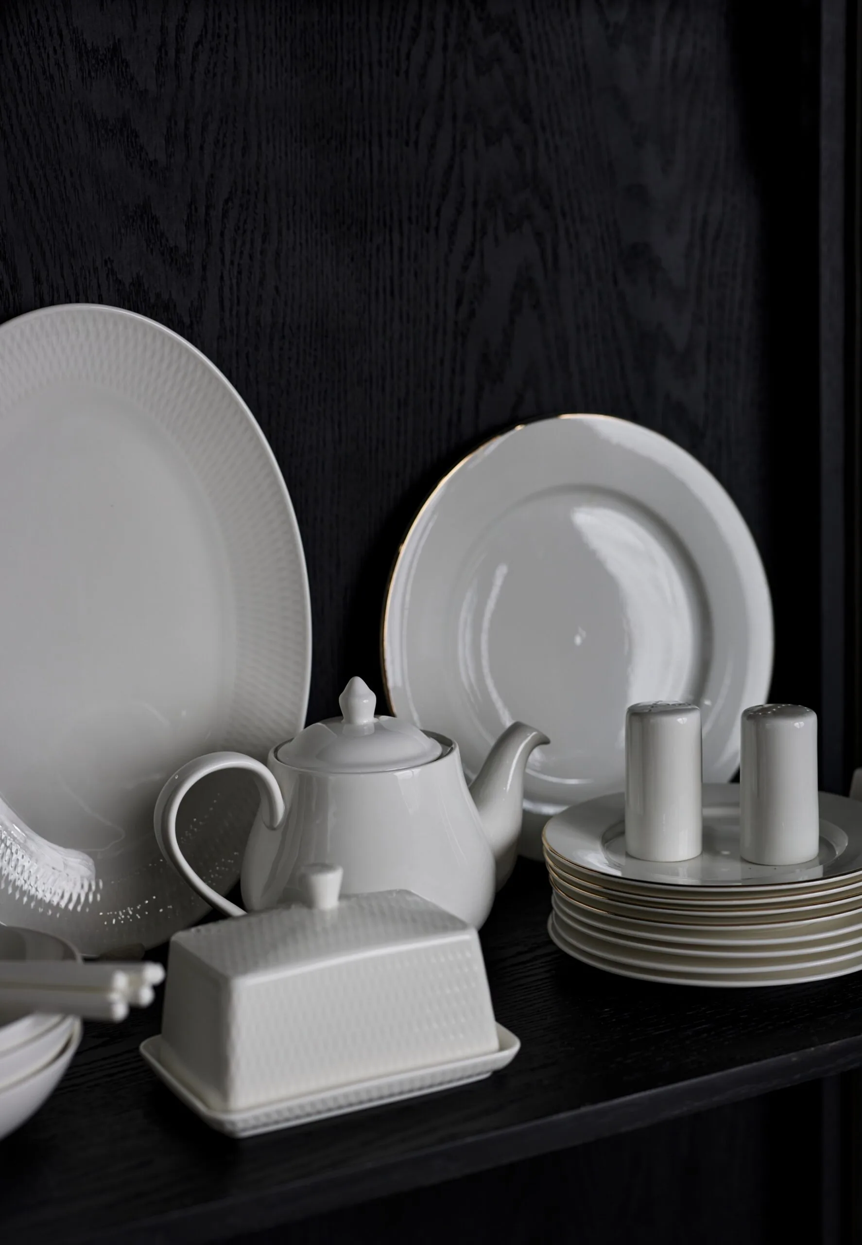 A black cabinet shelf with two round white plates, a white teapot, a white butter dish and a white stack of small bread plates are all displayed in the cabinet shelf.