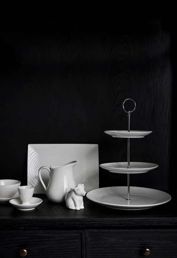 A black backdrop. A tiered white cake stand is in front, with a white milk jug, white rectangular platter and white egg holder with dish underneath.