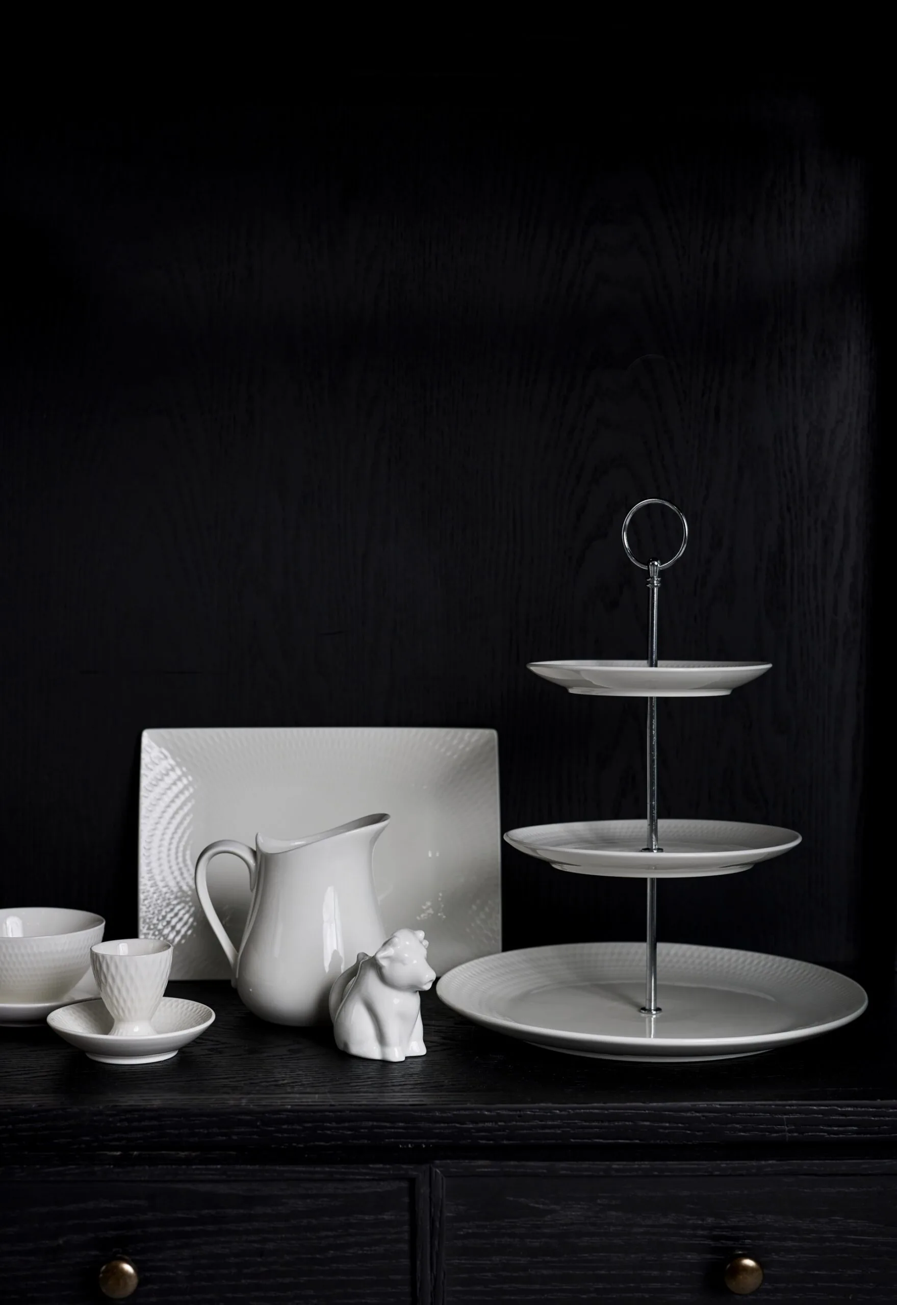 A black backdrop. A tiered white cake stand is in front, with a white milk jug, white rectangular platter and white egg holder with dish underneath.