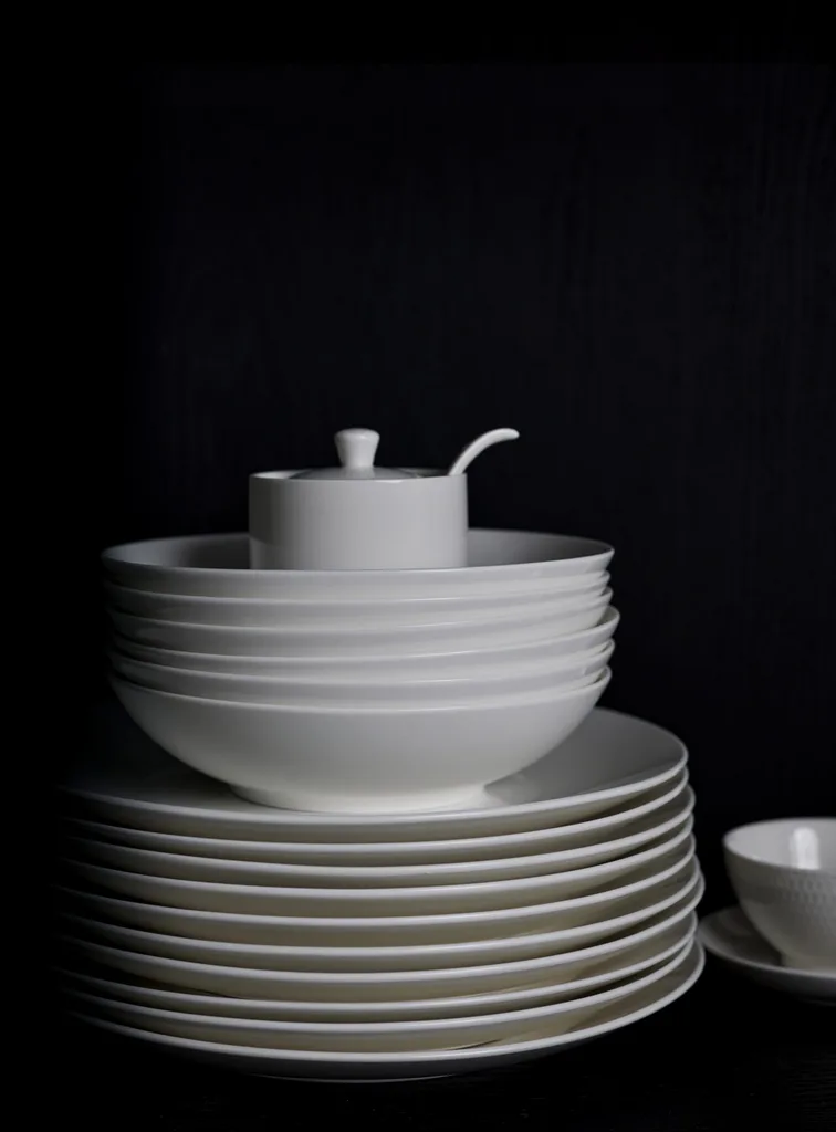 A stack of white plates with a stack of white bowls on top. A small white sugar dish is on top. The background is black.