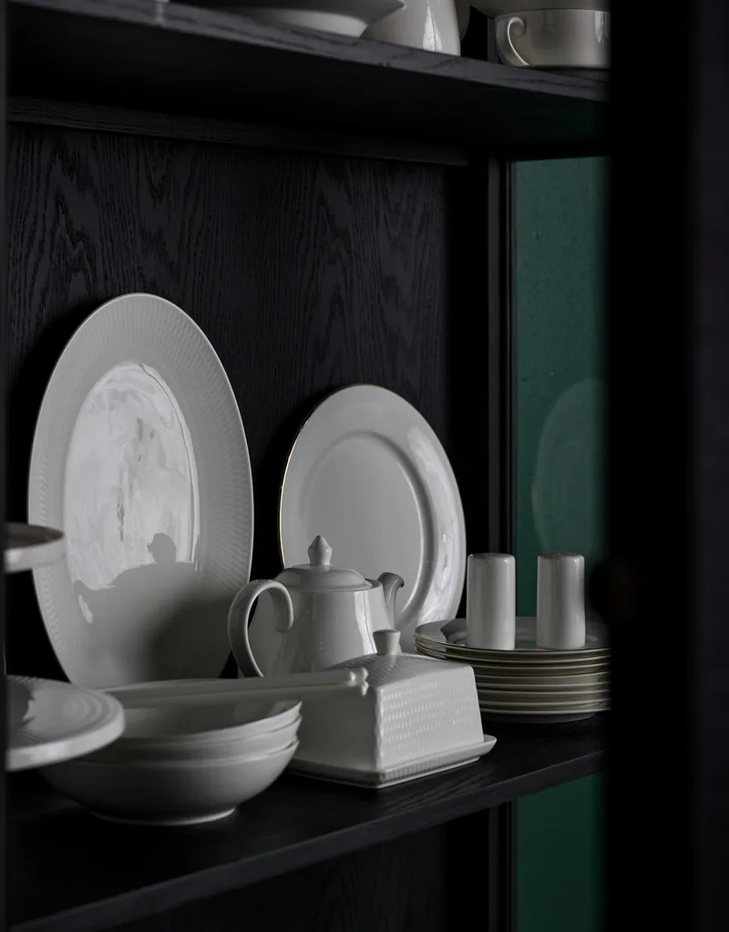 A black cabinet shelf with two round white plates, a white teapot, a white butter dish and a white stack of small bread plates are all displayed in the cabinet shelf.