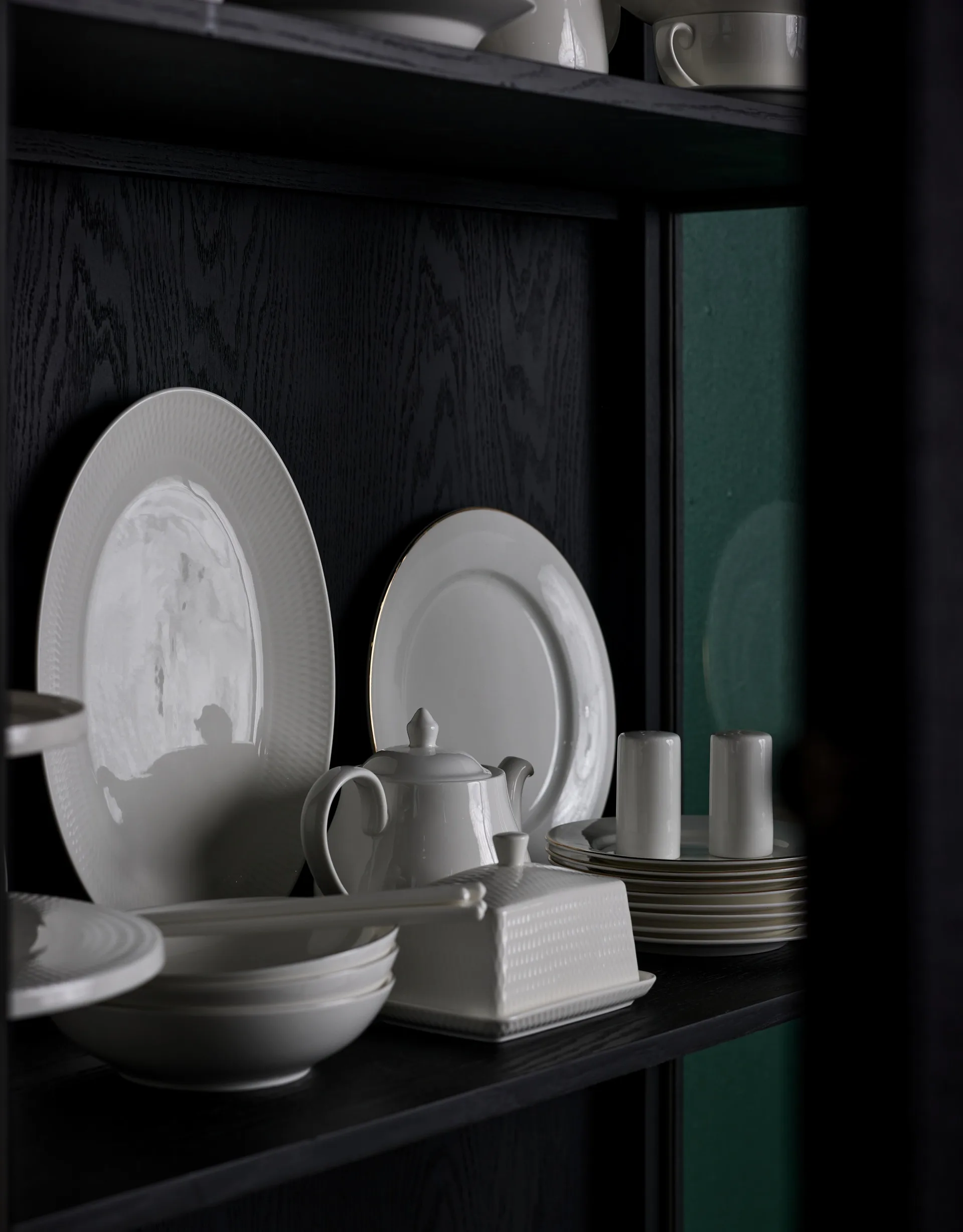A black cabinet shelf with two round white plates, a white teapot, a white butter dish and a white stack of small bread plates are all displayed in the cabinet shelf.