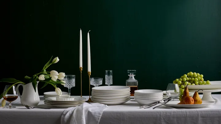 A white table in front of a plain dark backdrop. A white tablecloth drapes over the table, with stacks of white plates and bowls over the top. One plate has three pears in it, one white jug has white tulips in it. One white cake stand has a bunch of grapes on it. Two candlesticks are in brass candle holders.