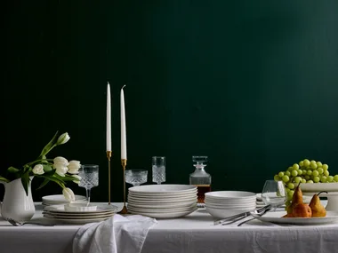 A white table in front of a plain dark backdrop. A white tablecloth drapes over the table, with stacks of white plates and bowls over the top. One plate has three pears in it, one white jug has white tulips in it. One white cake stand has a bunch of grapes on it. Two candlesticks are in brass candle holders.