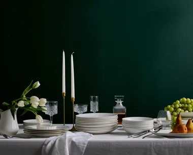A white table in front of a plain dark backdrop. A white tablecloth drapes over the table, with stacks of white plates and bowls over the top. One plate has three pears in it, one white jug has white tulips in it. One white cake stand has a bunch of grapes on it. Two candlesticks are in brass candle holders.