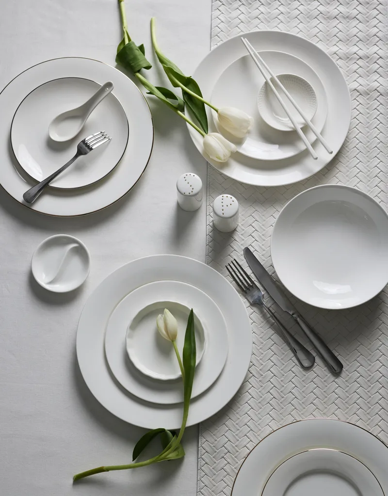 A white tablecloth background. Three stacks of plates are on the table, beside white tulips. Silver cutlery and two white ceramic chopsticks are also on the table.