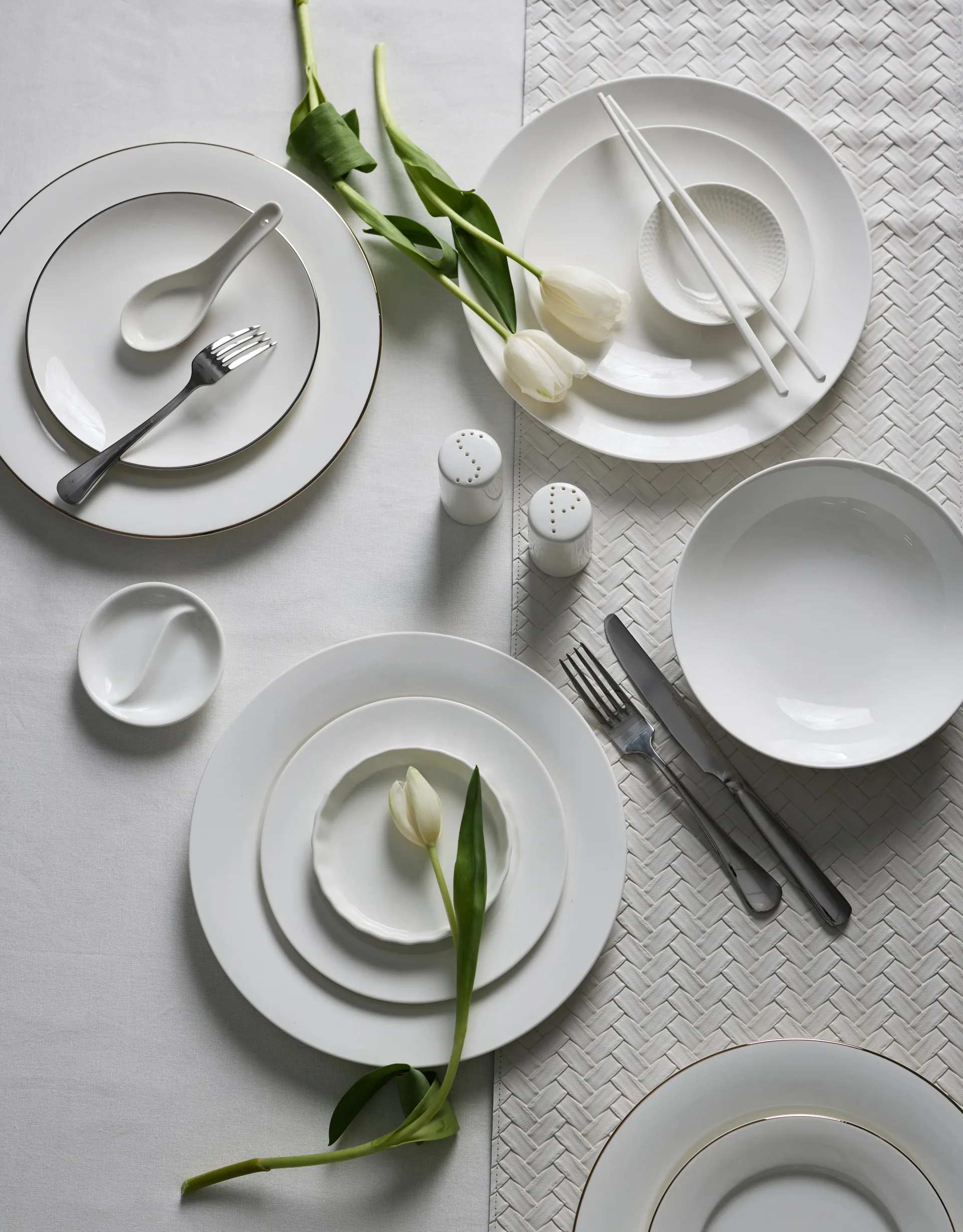 A white tablecloth background. Three stacks of plates are on the table, beside white tulips. Silver cutlery and two white ceramic chopsticks are also on the table.