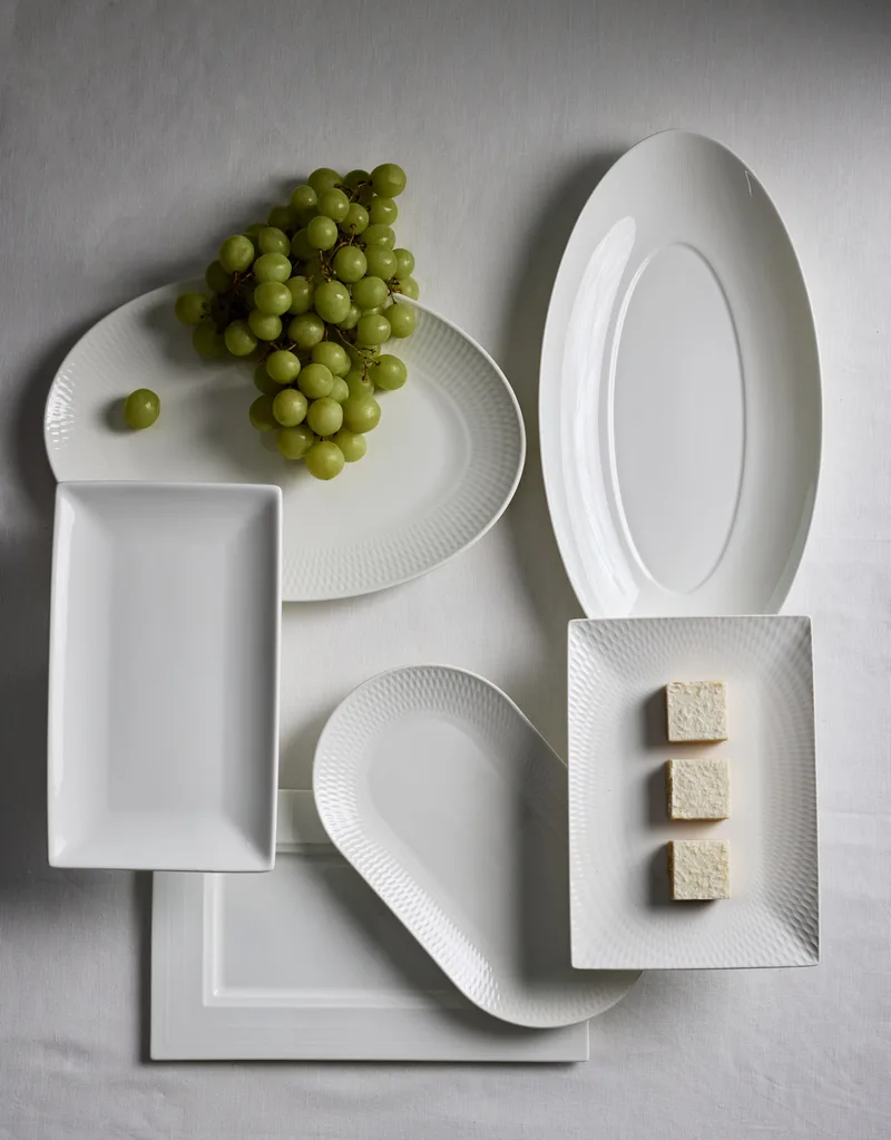 A white tablecloth based with a mix of white platters on top. Some are oval, some are rectangular and some are round. A bunch of green grapes and three small squares of cake are on two of the platters.