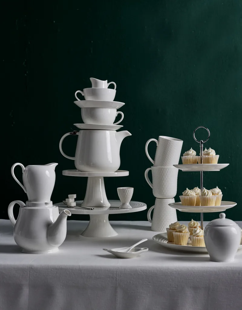 A white table in front of a plain dark backdrop. A white tablecloth drapes over the table, with white teapots and cake stands standing on top of one another. A tiered cupcake stand is to the side, beside a stack of white mugs, and is decorated with vanilla cupcakes.