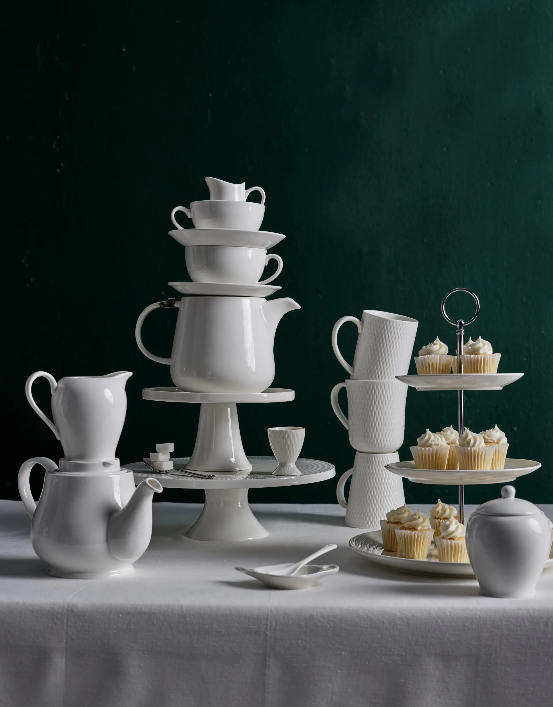 A white table in front of a plain dark backdrop. A white tablecloth drapes over the table, with white teapots and cake stands standing on top of one another. A tiered cupcake stand is to the side, beside a stack of white mugs, and is decorated with vanilla cupcakes.