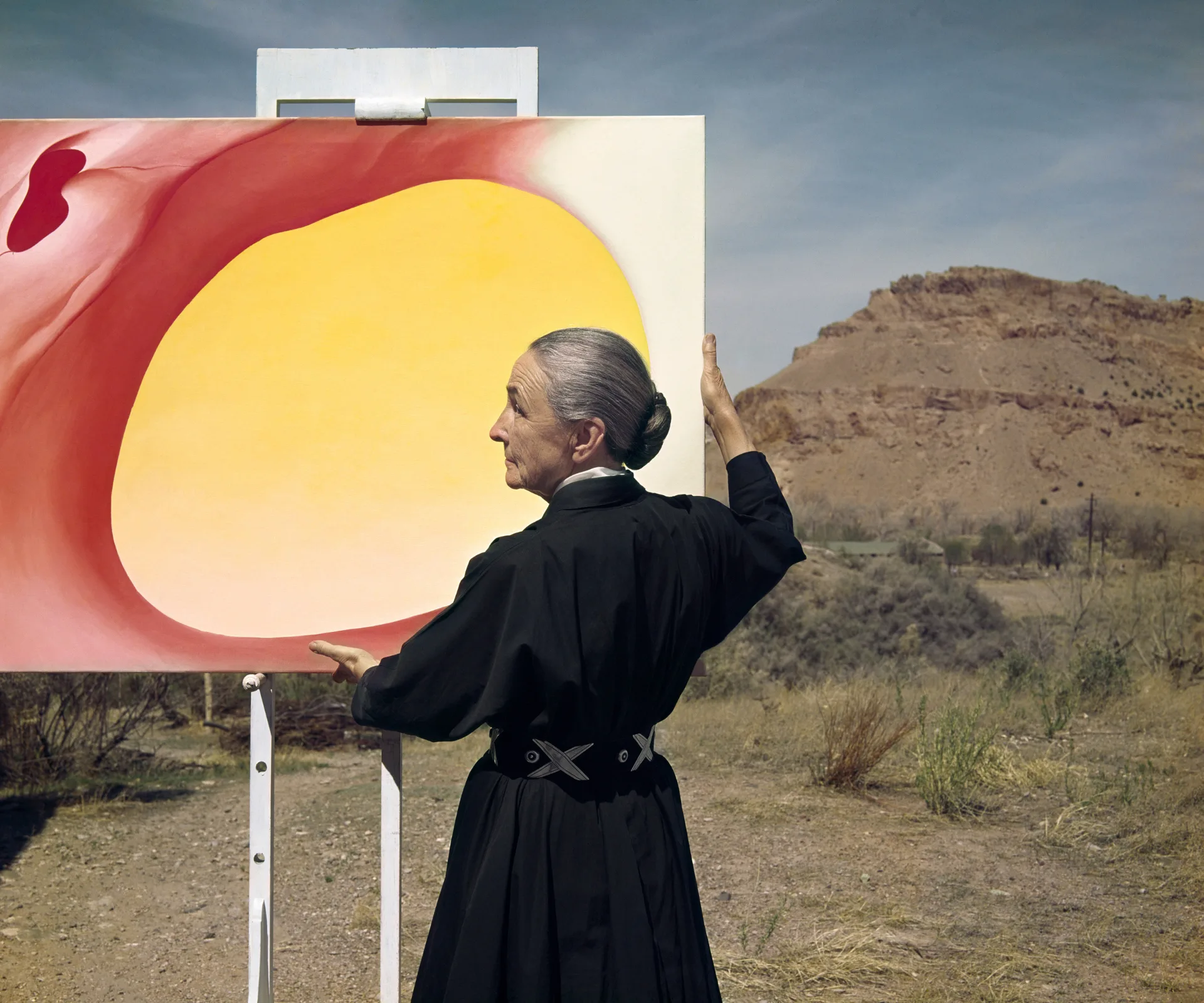 Georgia O'Keeffe Painter ABIQUIU, NEW MEXICO : 1960 - The American painter Georgia O'Keeffe is standing outside her art studio holing her pelvis series color painting that has center yellow and edges red. She si holding on to the edges of her painting looking sideways.