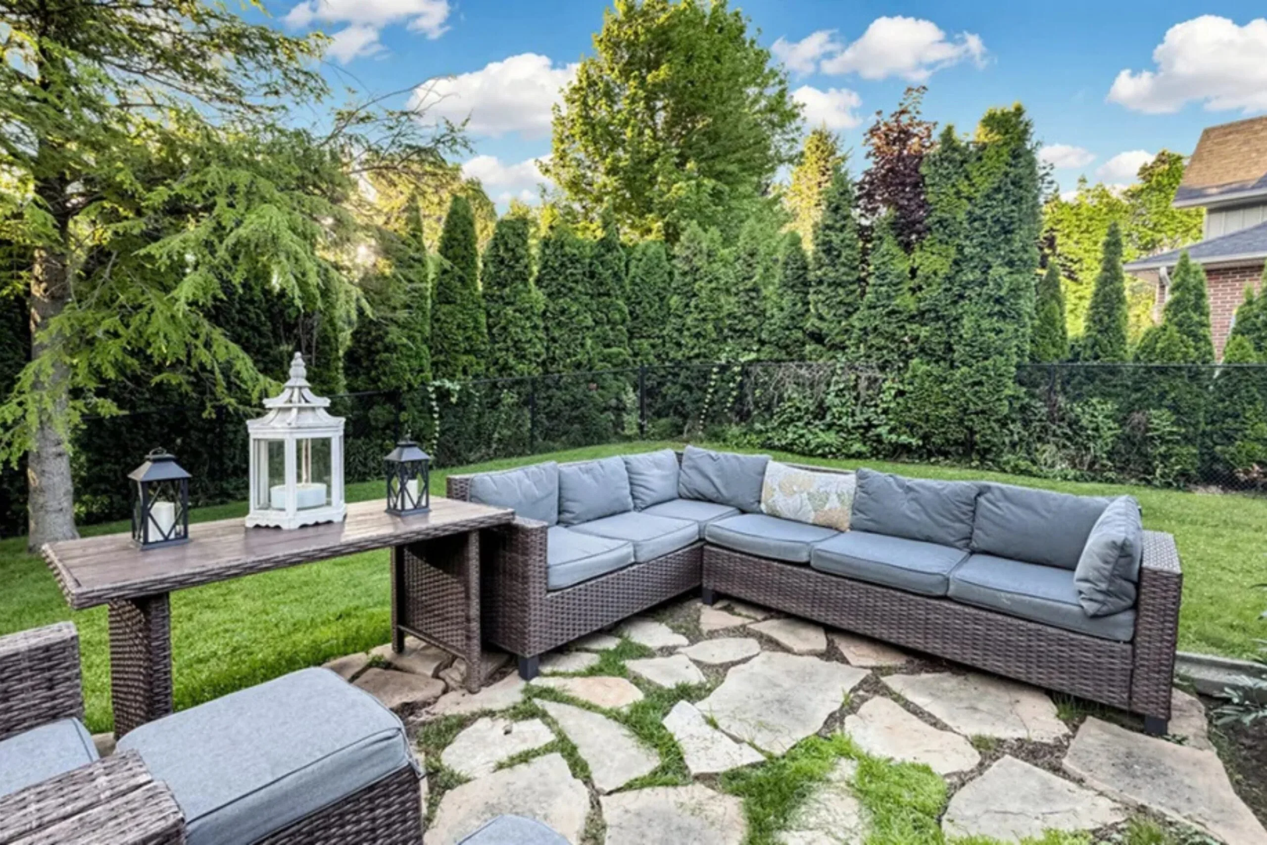 Stone-paved outdoor entertaining area with wicker lounge furniture set among mature trees and manicured gardens.