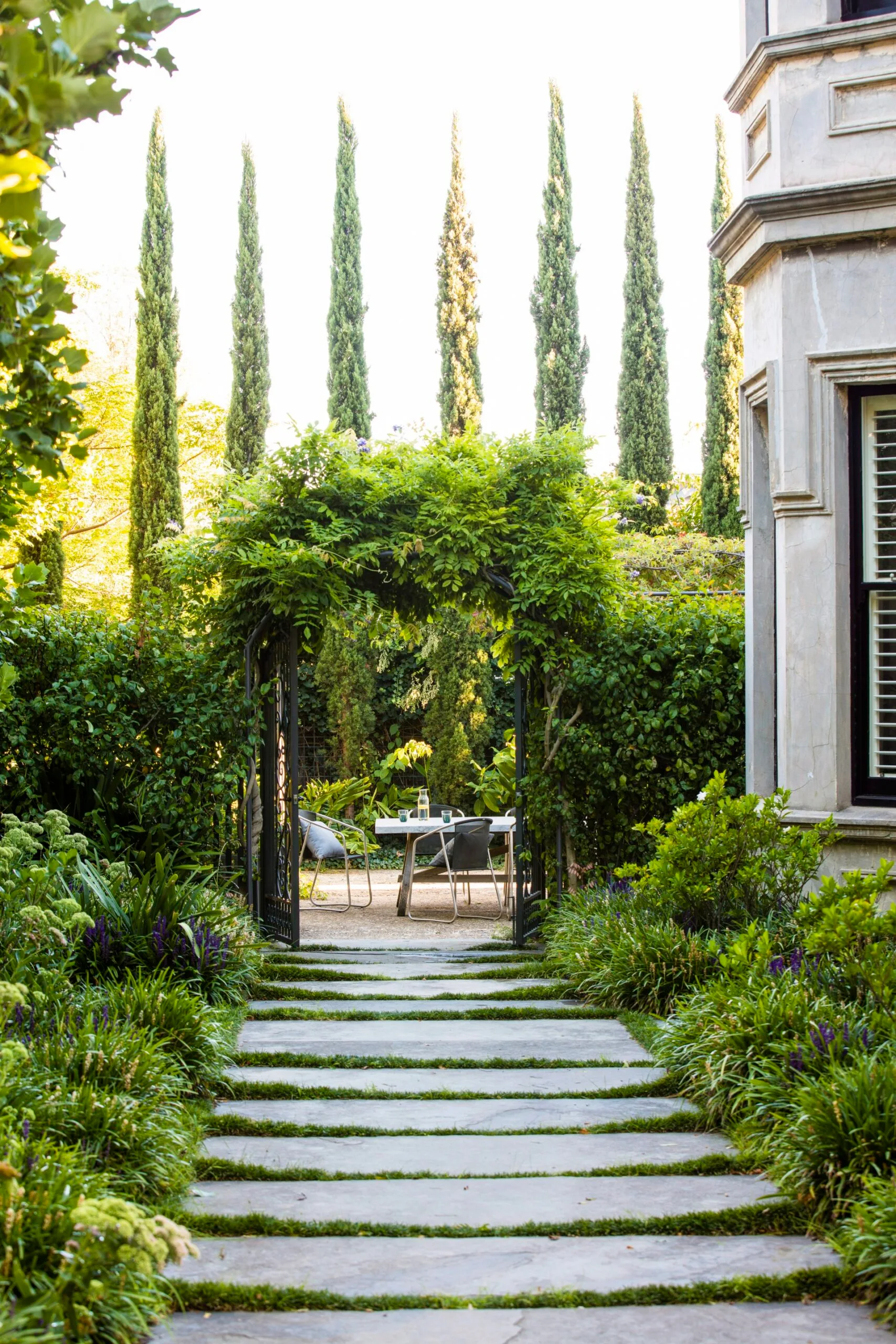 Stone path leads to a lush garden with gateway that is covered in greenery. Beyond the open gate is an outdoor dining area, surrounded by tall cypress trees and greenery. Soft forms of Liriope muscari, Sedum and gardenia create informal hedging.