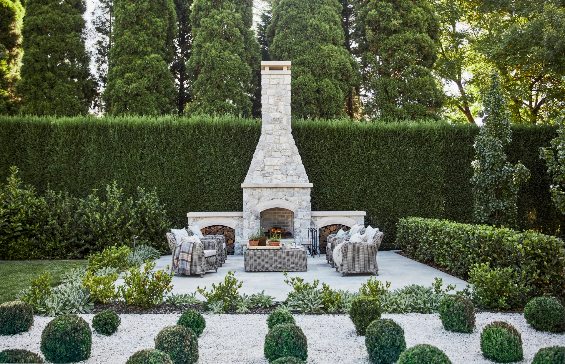 Outdoor stone fireplace with wicker chairs and lush greenery surrounding a manicured garden. In the foreground, neatly trimmed Buxus balls form low-lying topiaries while in the background a large hedge is positioned behind the fireplace.