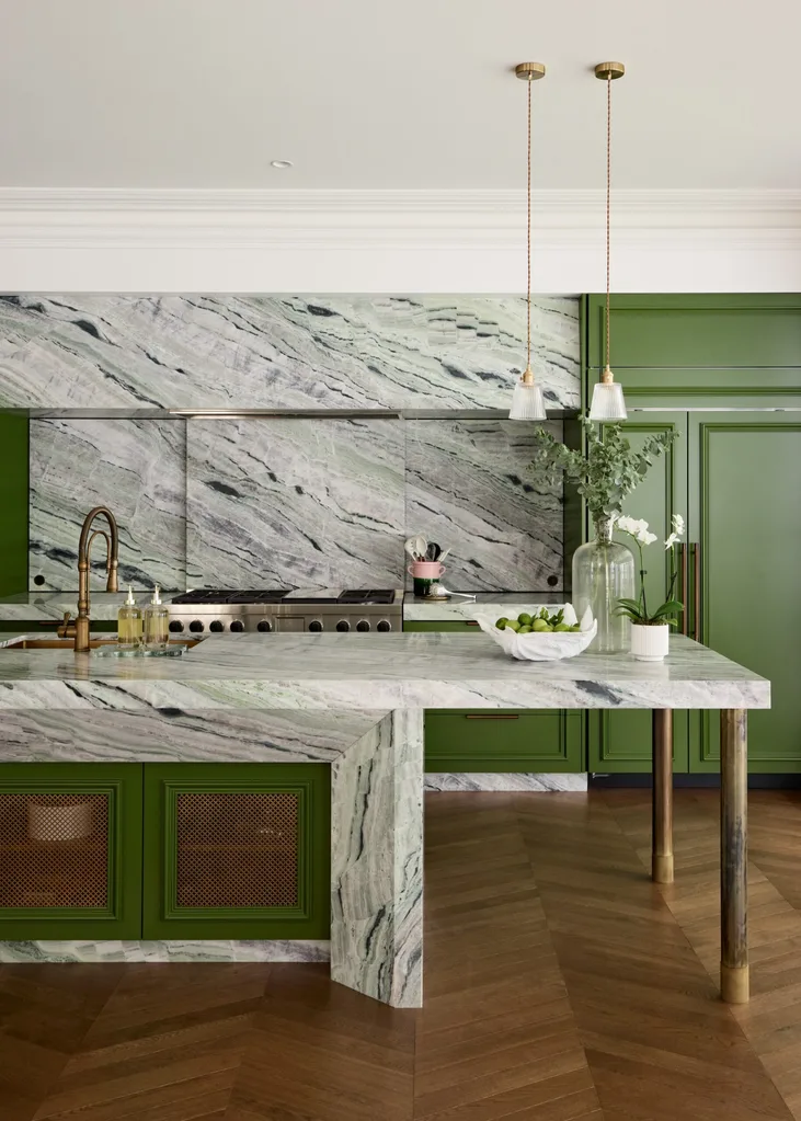 A large family kitchen that balances the latest kitchen trends with a sense of timelessness. Boldly veined stone covers the benchtops, splashbacks, rangehood cover and island surrounds. A deep green paint has been used on the joinery profiles, while brass pendants hang from the ceiling. Brass mesh squares are on the joinery profiles on the side of the kitchen island.