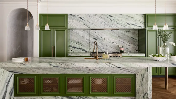 A large family kitchen that balances the latest kitchen trends with a sense of timelessness. Boldly veined stone covers the benchtops, splashbacks, rangehood cover and island surrounds. A deep green paint has been used on the joinery profiles, while brass pendants hang from the ceiling. Brass mesh squares are on the joinery profiles on the side of the kitchen island.
