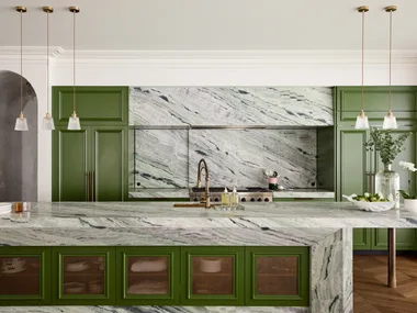 A large family kitchen that balances the latest kitchen trends with a sense of timelessness. Boldly veined stone covers the benchtops, splashbacks, rangehood cover and island surrounds. A deep green paint has been used on the joinery profiles, while brass pendants hang from the ceiling. Brass mesh squares are on the joinery profiles on the side of the kitchen island.