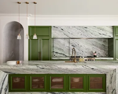 A large family kitchen that balances the latest kitchen trends with a sense of timelessness. Boldly veined stone covers the benchtops, splashbacks, rangehood cover and island surrounds. A deep green paint has been used on the joinery profiles, while brass pendants hang from the ceiling. Brass mesh squares are on the joinery profiles on the side of the kitchen island.