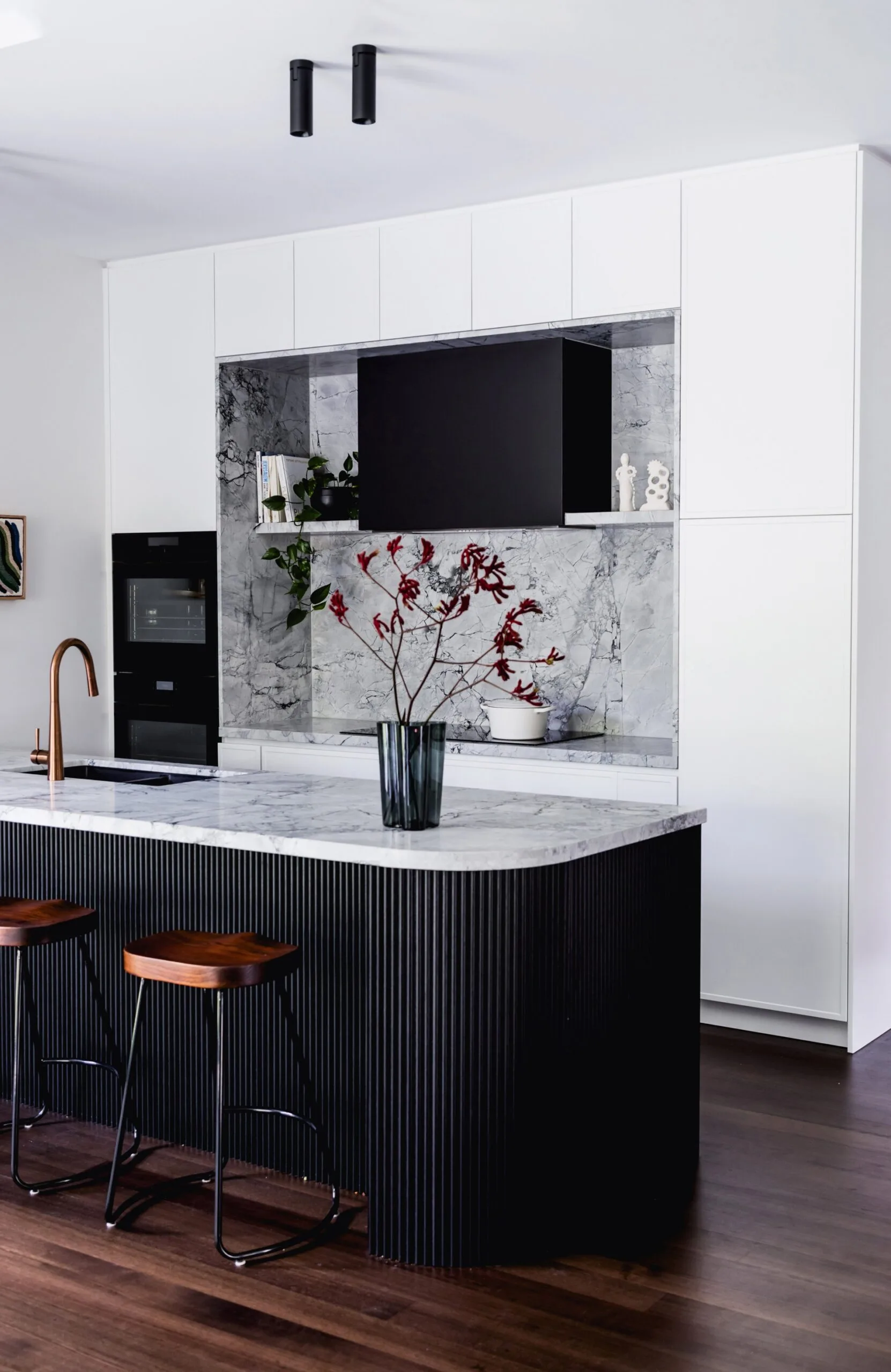 A contemporary kitchen in a heritage home that balances modern style and traditional architectural details to create a sense of timelessness. The rangehood is in an alcove, with a boldly veined marble on the benchtops, sides and splashback wall. The rangehood cover is black, as is the surrounds of the kitchen island. White joinery surrounds the rangehood alcove.