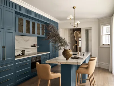 A large family kitchen that balances the latest kitchen trends with a sense of timelessness. The joinery profiles are finished in a shade of blue that is a bold contrast to the white ceilings, white marble benchtops and splashback and also the light toned timber flooring. Brass tapware is on the sink (built into the kitchen island) and a pendant light above the island is brass. The kitchen island has three light brown bar stools surrounding it.