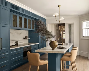 A large family kitchen that balances the latest kitchen trends with a sense of timelessness. The joinery profiles are finished in a shade of blue that is a bold contrast to the white ceilings, white marble benchtops and splashback and also the light toned timber flooring. Brass tapware is on the sink (built into the kitchen island) and a pendant light above the island is brass. The kitchen island has three light brown bar stools surrounding it.
