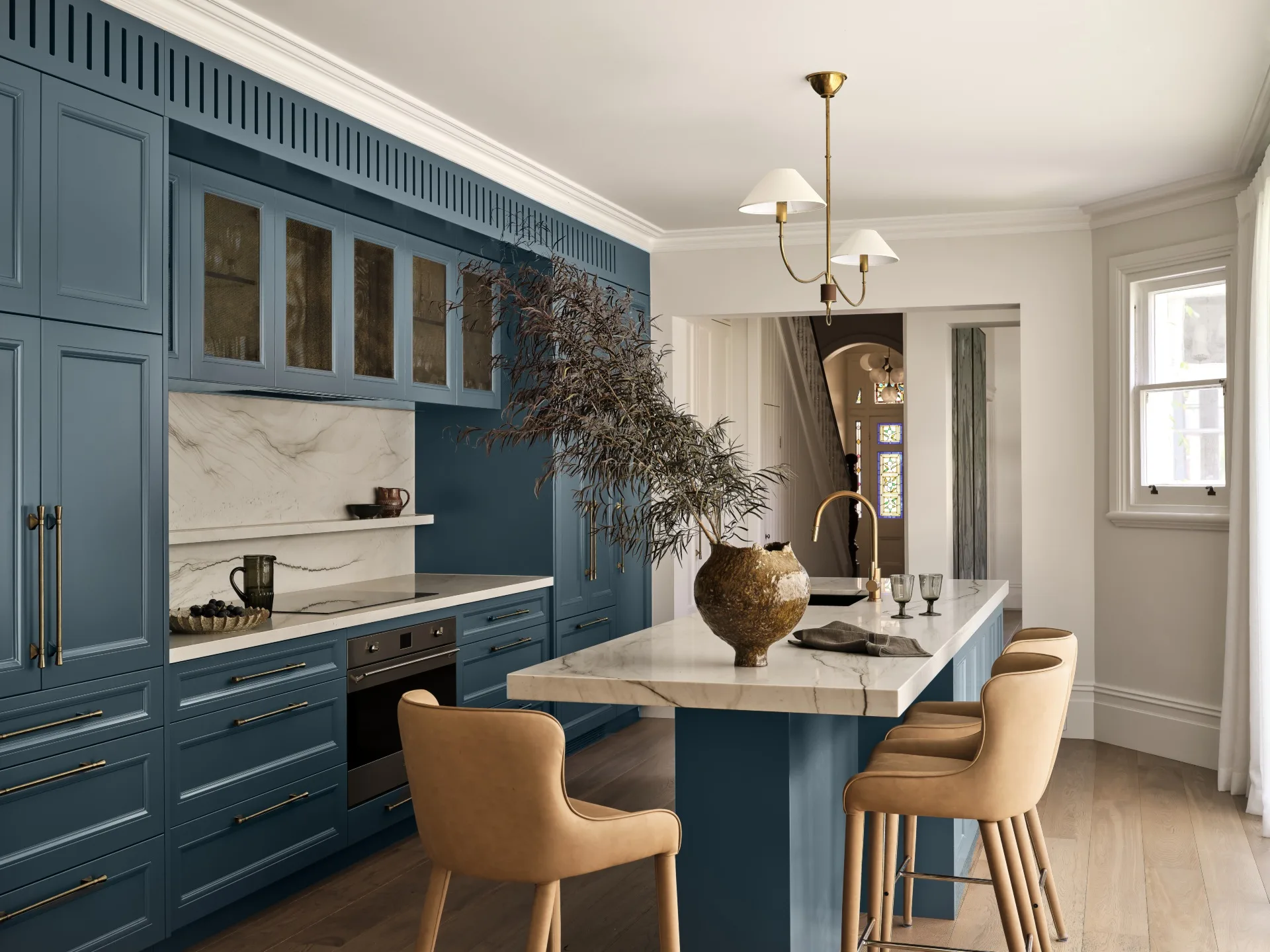 A large family kitchen that balances the latest kitchen trends with a sense of timelessness. The joinery profiles are finished in a shade of blue that is a bold contrast to the white ceilings, white marble benchtops and splashback and also the light toned timber flooring. Brass tapware is on the sink (built into the kitchen island) and a pendant light above the island is brass. The kitchen island has three light brown bar stools surrounding it.