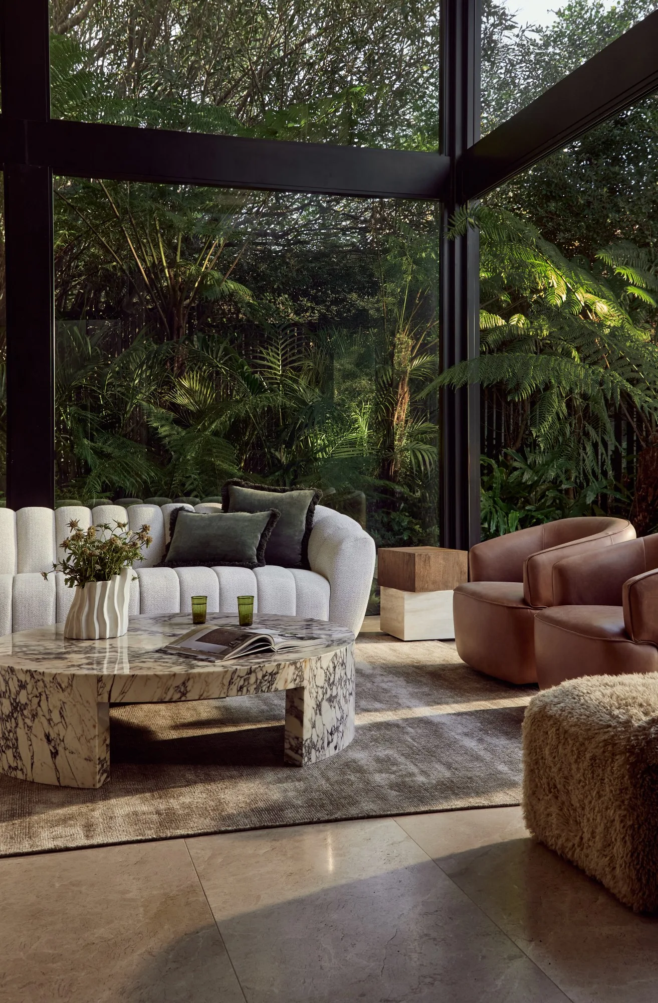Elegant living room with marble table, beige sofa, brown chairs, large windows, and lush greenery outside.